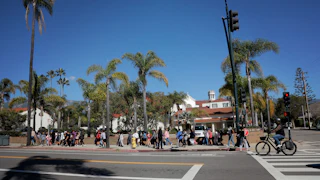 A view of a Santa Barbara California city street, with people walking on a sidewalk in front of palm treets and buildings.