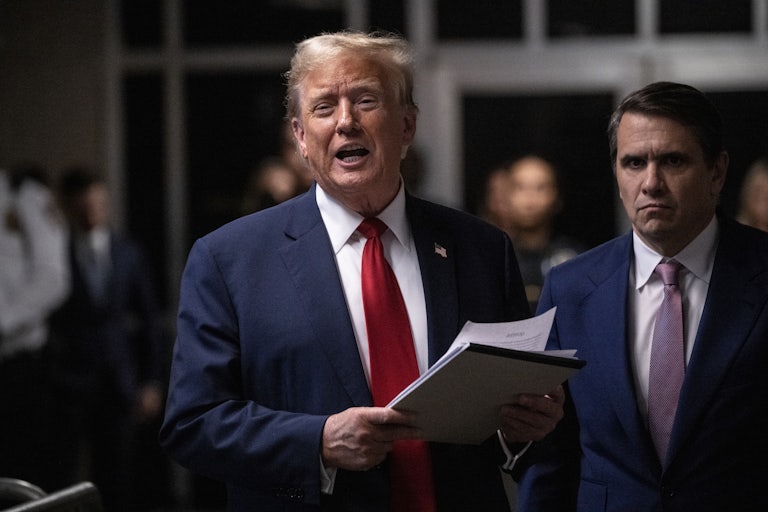 Donald Trump in the courtroom holds a stack of papers and speaks, brows furrowed. His lawyer Todd Blanche stands beside him, also brows furrowed, staring at the camera.