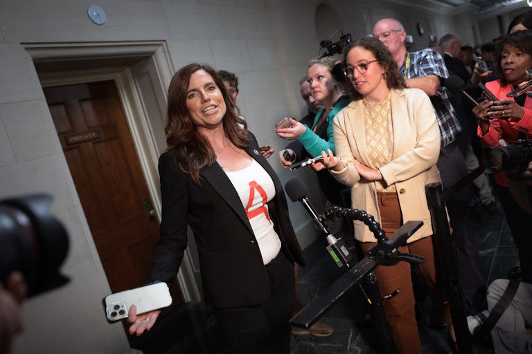 Nancy Mace wears a black blazer and a white shirt with a red "A" emblazoned on it, as reporters and mics surround her.