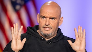 John Fetterman holds his hands up while speaking in front of an American flag. He's wearing a hoodie