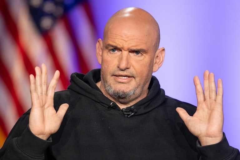 John Fetterman holds his hands up while speaking in front of an American flag. He's wearing a hoodie
