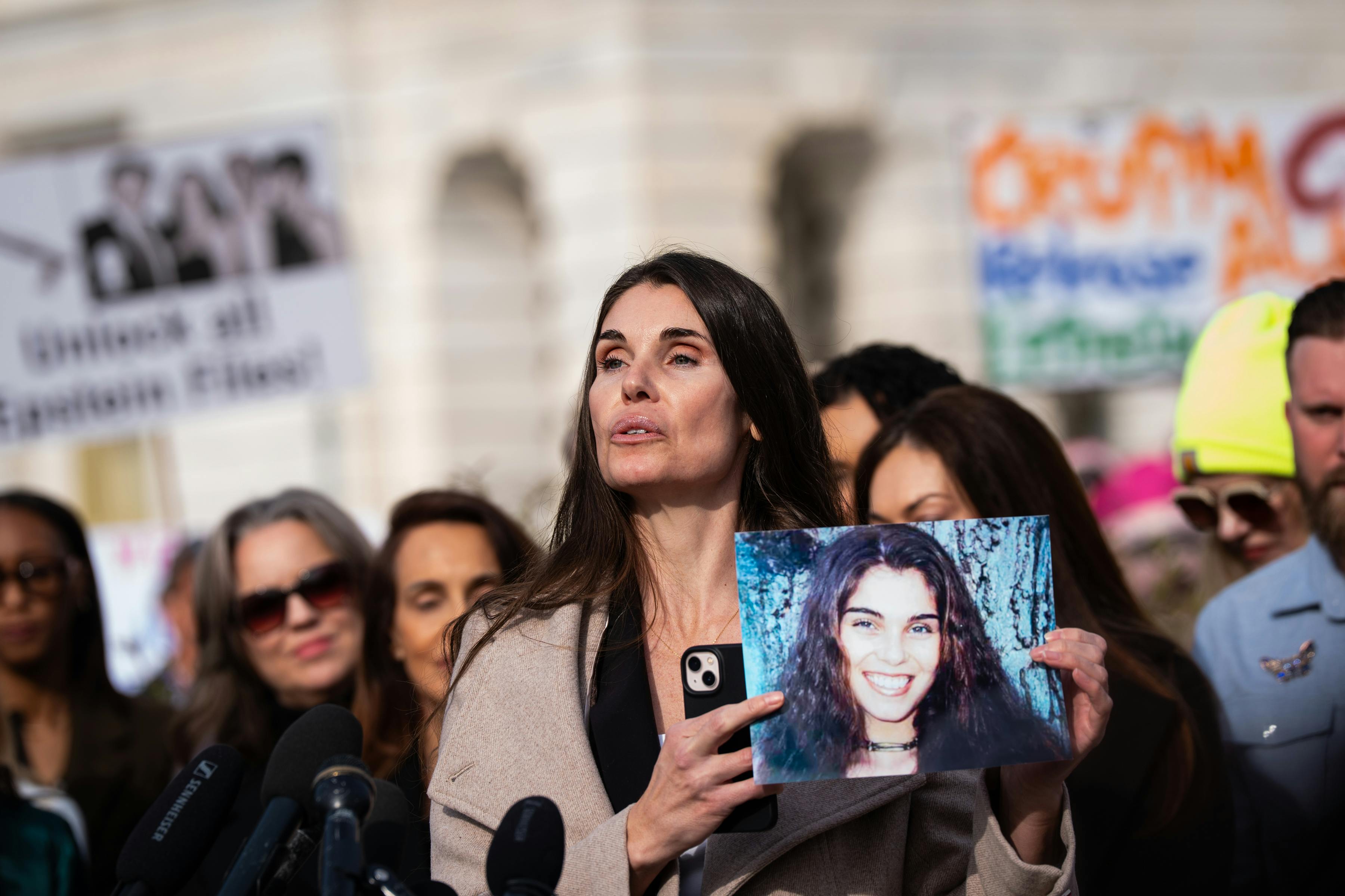 An Epstein survivor holds a picture of herself up in a press conference outside the Capitol. Others gather behind her.
