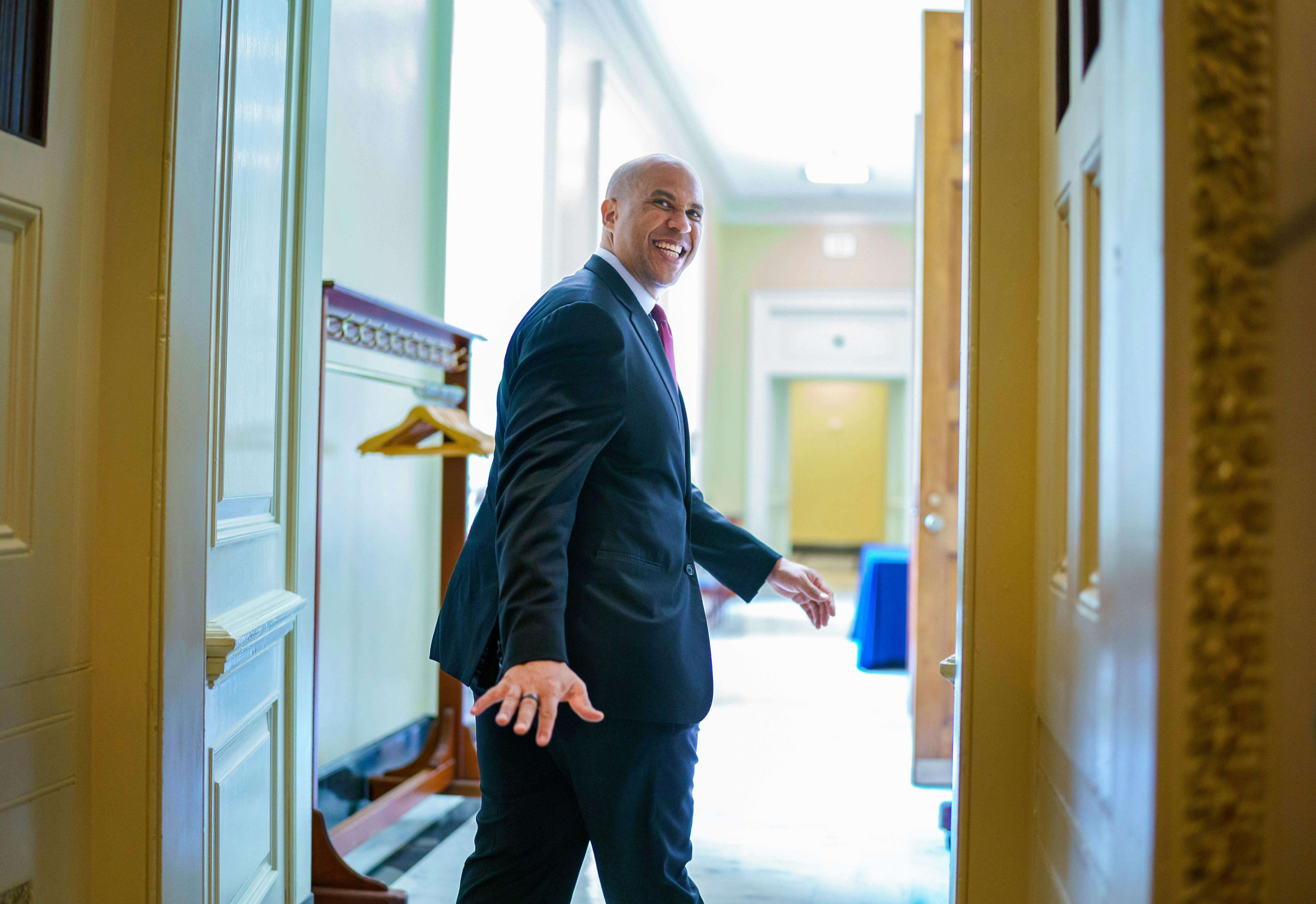 A smiling Senator Cory Booker looks back to wave goodbye as he leaves a room.