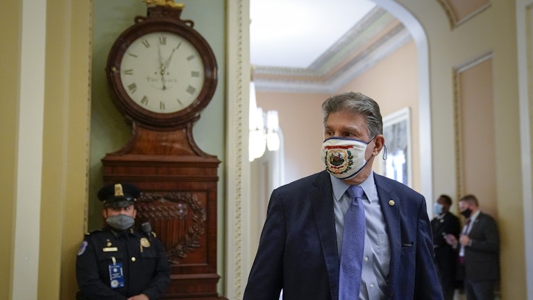 Clad in a mask with the West Virginia state flag on it, Senator Joe Manchin walks down a Capitol hallway in front of a masked guard.