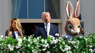 Donald Trump speaks at a microphone on a balcony. Melania Trump stands to his right and someone in an Easter Bunny costume stands to his left.