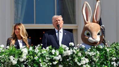 Donald Trump speaks at a microphone on a balcony. Melania Trump stands to his right and someone in an Easter Bunny costume stands to his left.