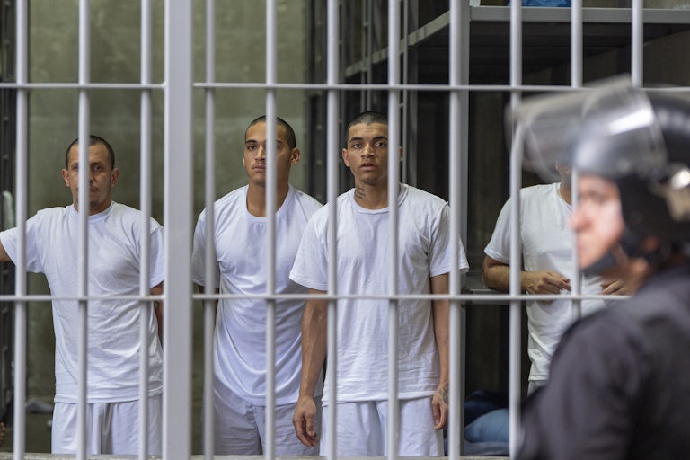 Three men wearing white T-shirts and shorts look out behind the bars in their cell in El Salvador’s megaprison.