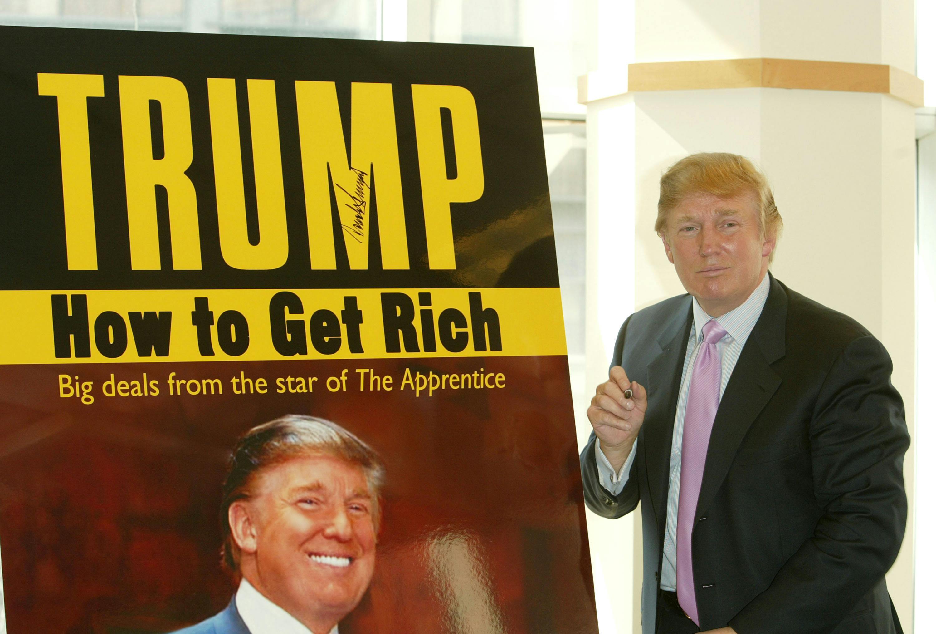 Donald Trump stands in front of a poster advertising one of his books.