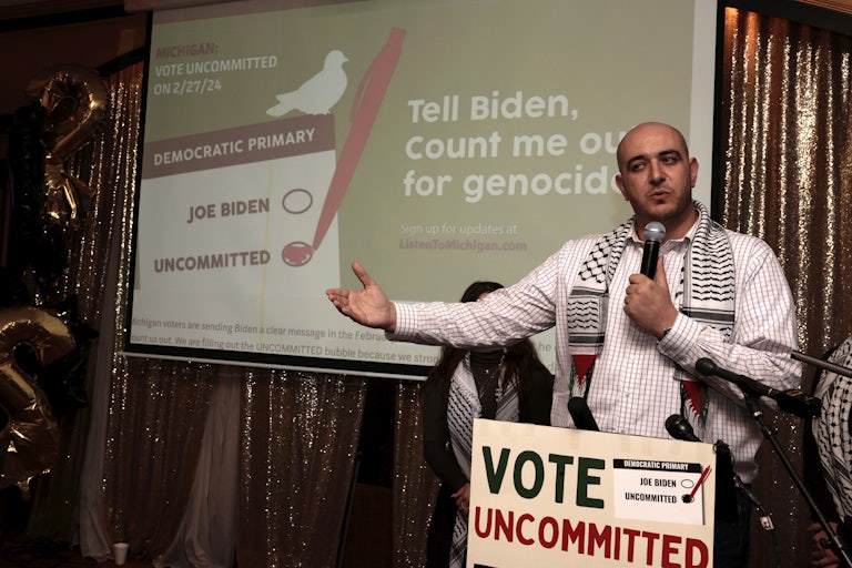 A man wearing a dress shirt and a keffiyeh speaks behind a sign reading "Vote uncommitted"