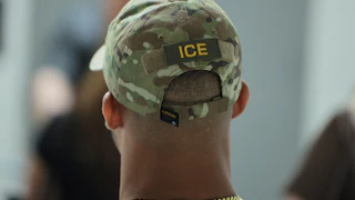 ICE agent (back of their head, wearing a camo ICE cap and a thick gold chain)