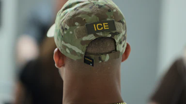 ICE agent (back of their head, wearing a camo ICE cap and a thick gold chain)