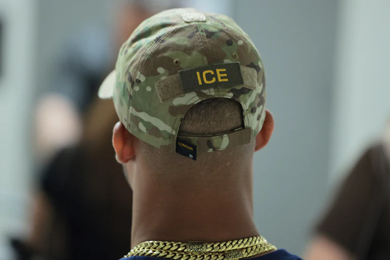 ICE agent (back of their head, wearing a camo ICE cap and a thick gold chain)