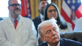 Donald Trump's eyes droop as he sits at his desk in the Oval Office of the White House. People in white lab coats stand behind him.