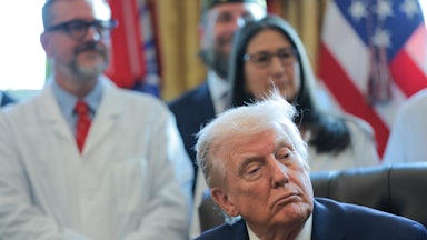 Donald Trump's eyes droop as he sits at his desk in the Oval Office of the White House. People in white lab coats stand behind him.