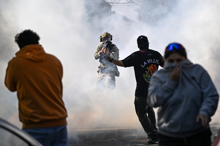 A masked federal agent points a gun at someone as tear gas floats in the air and others try to escape.
