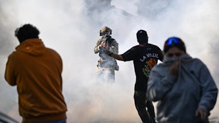 A masked federal agent points a gun at someone as tear gas floats in the air and others try to escape.
