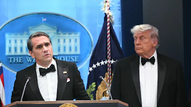 Acting Attorney General Todd Blanche speaks at a podium while Donald Trump stands next to him