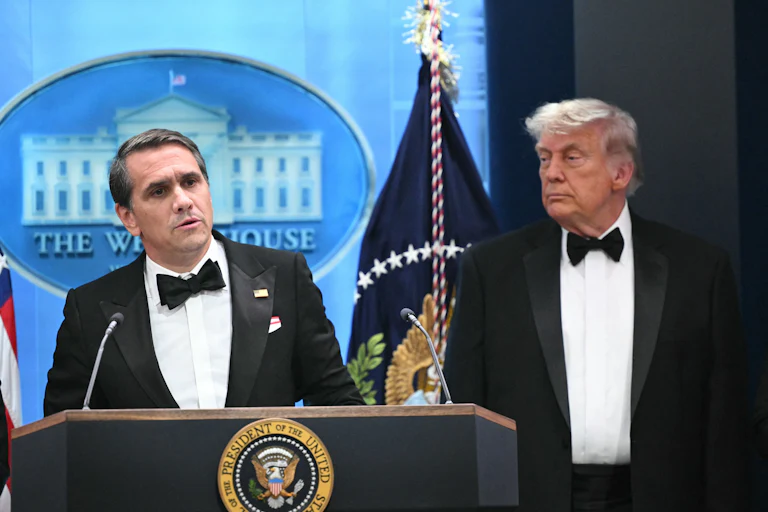 Acting Attorney General Todd Blanche speaks at a podium while Donald Trump stands next to him