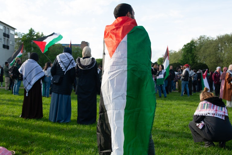 A student (back to the camera) is draped in a Palestinian flag. Others are in the background wearing keffiyehs and a Palestinian flag.