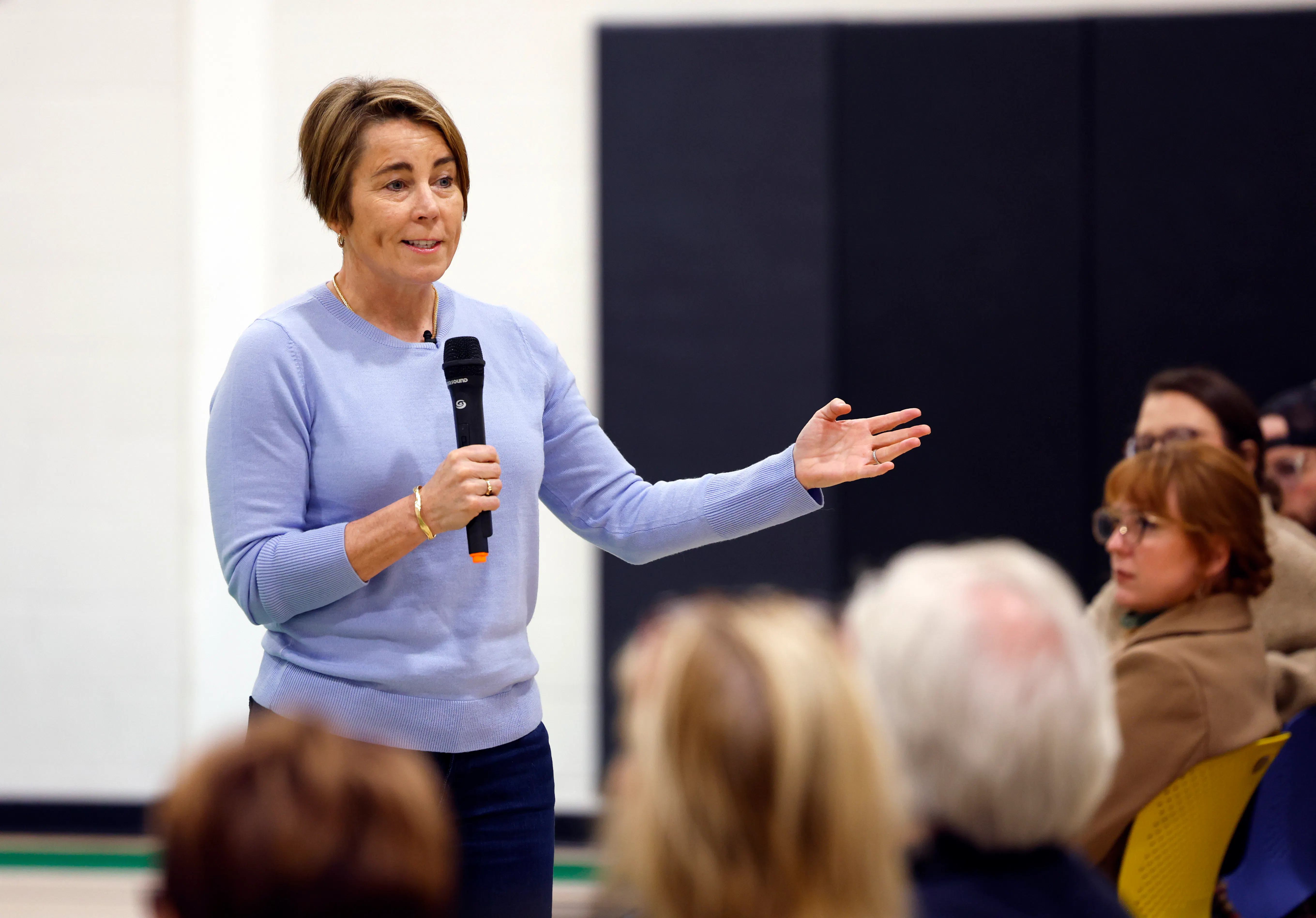 Maura Healey gestures while speaking into a microphone, in front of a row of seated people.