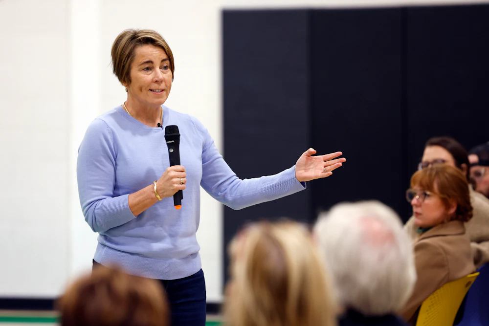 Maura Healey gestures while speaking into a microphone, in front of a row of seated people.