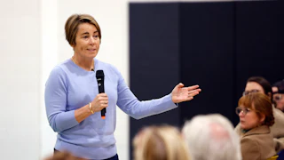 Maura Healey gestures while speaking into a microphone, in front of a row of seated people.