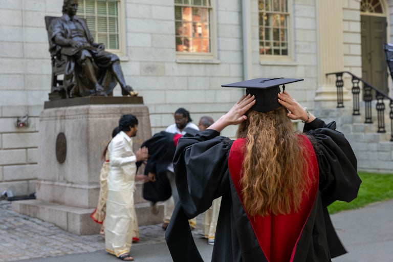 A student with long blond curly hair adjusts their gradation cap. Several Black and brown people are in the background.