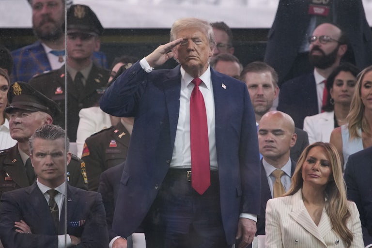 Donald Trump stands and salutes at the Army Parade, while standing behind a glass wall. Melania Trump and Pete Hegseth sit beside him, as do others.