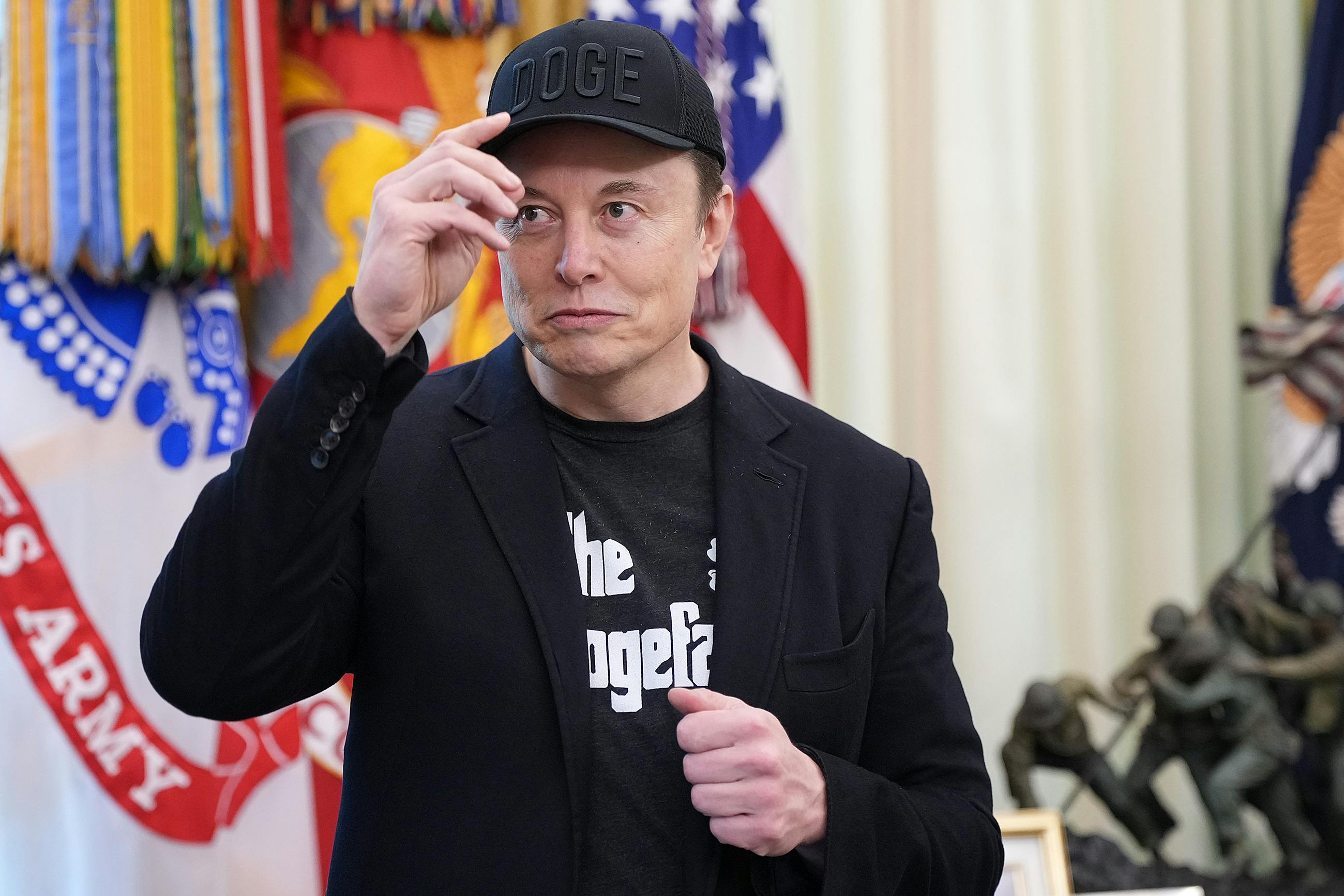 Elon Musk wears a DOGE cap and a shirt that reads "The Dogefather," in the same font as The Godfather movies.