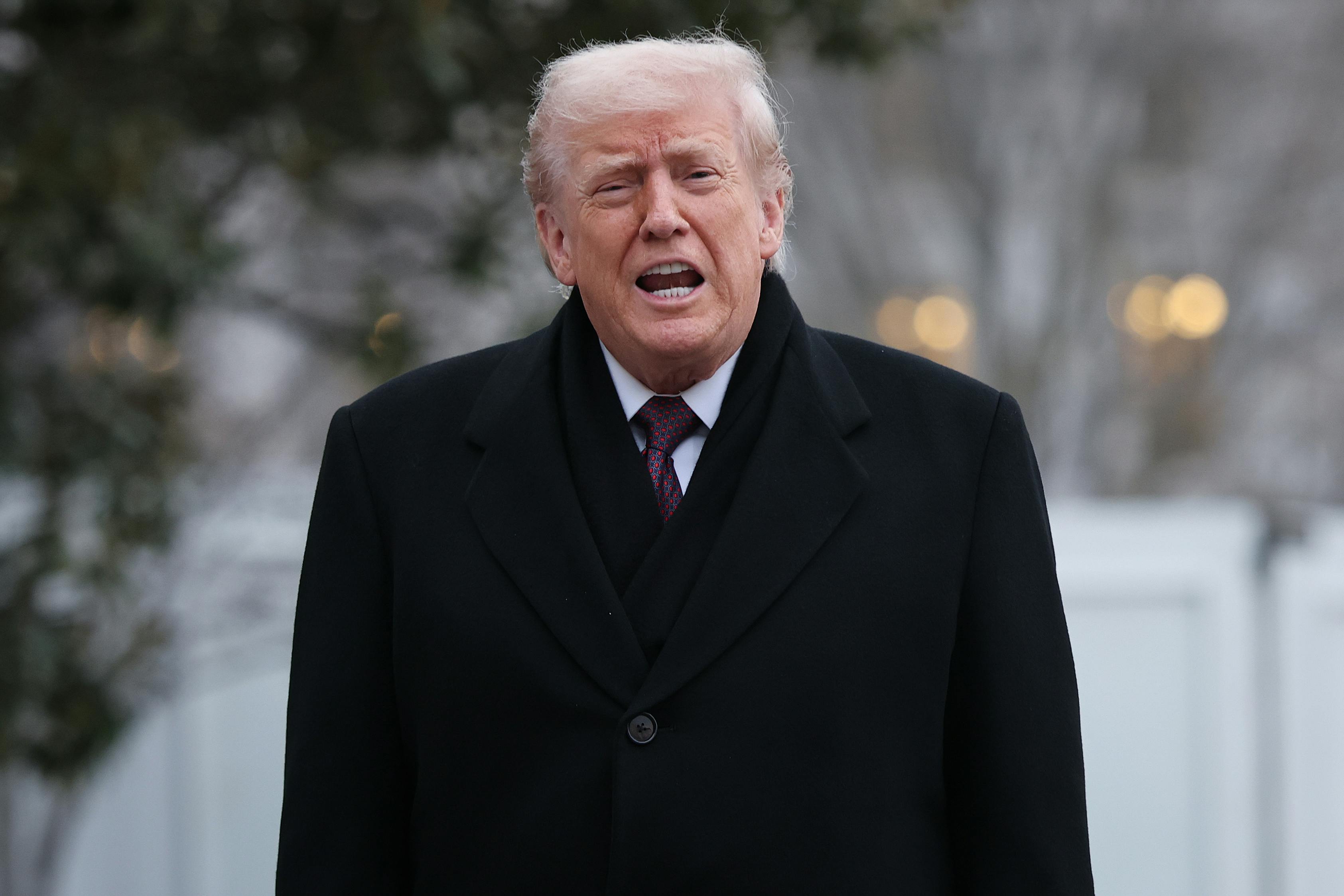 Trump opens his mouth to say something while standing outside in a dark overcoat.