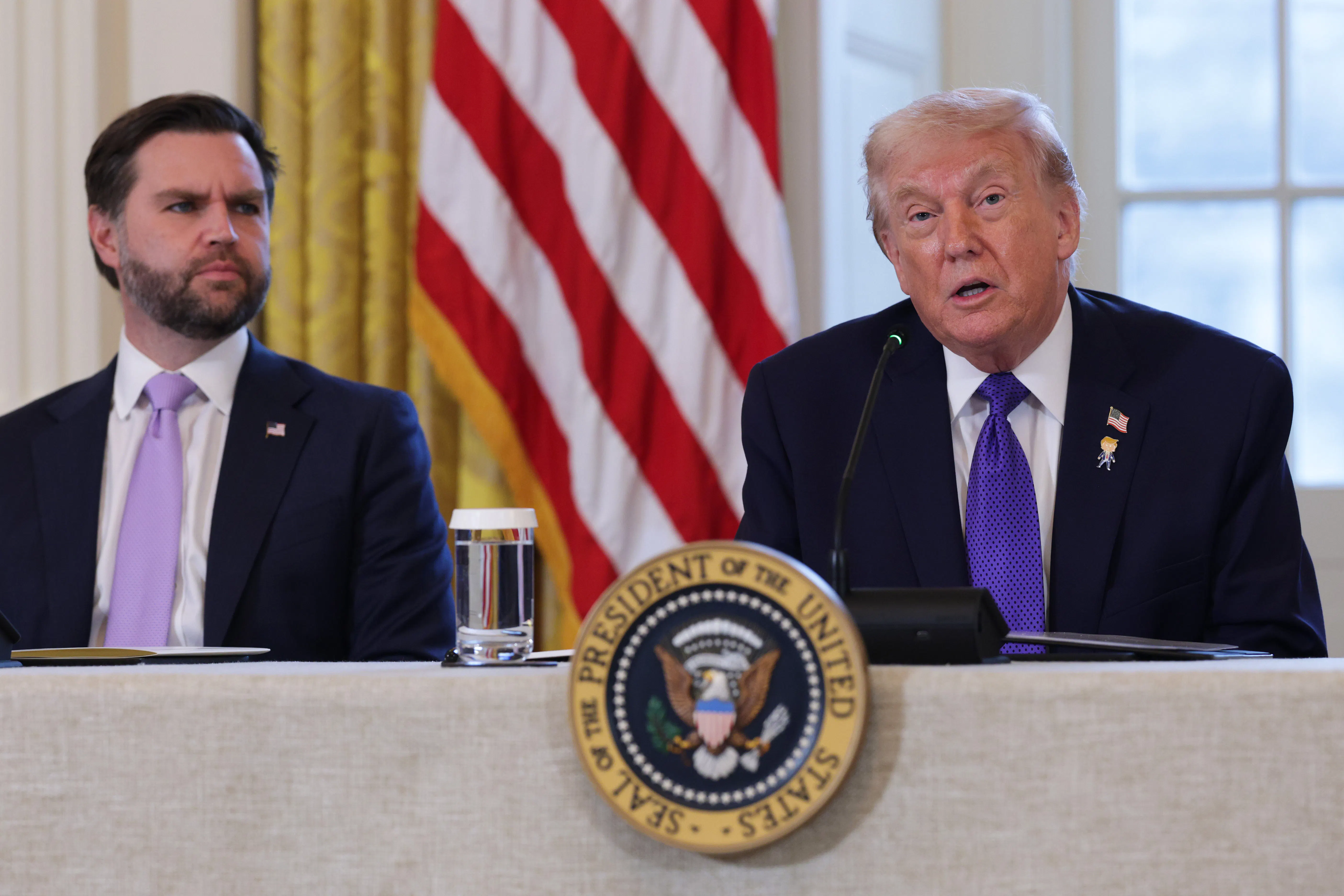 Donald Trump and JD Vance are both seated at a table with the presidential seal mounted to the front of the table. Trump is speaking, while Vance looks on to Trump's right. A glass of water is visible between Vance and Trump.