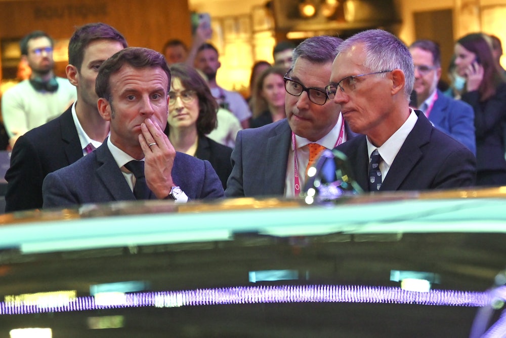 Carlos Tavares looks at Emanuel Macron, who frowns at a car.