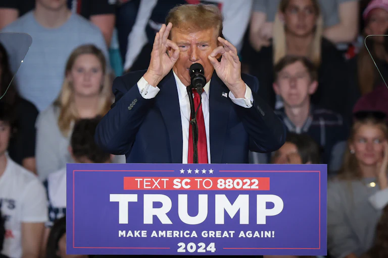 Donald Trump stands at a podium that reads "Text SC to 88022 Trump Make America Great Again 2024." He quints and makes an ok hand signal on both hands.
