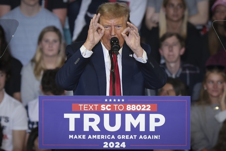 Donald Trump stands at a podium that reads "Text SC to 88022 Trump Make America Great Again 2024." He quints and makes an ok hand signal on both hands.