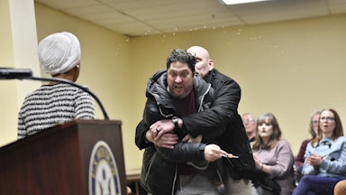 Security holds back a man with a syringe in his hand, as Ilhan Omar looks at them.