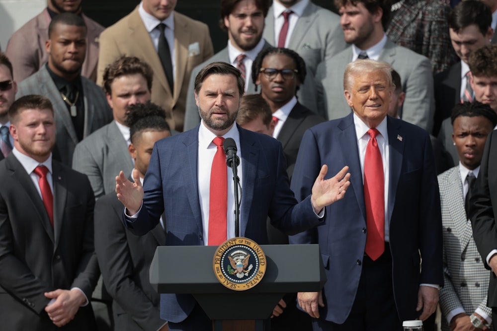 JD Vance speaks at a podium with Donald Trump beside him, flanked by other people.