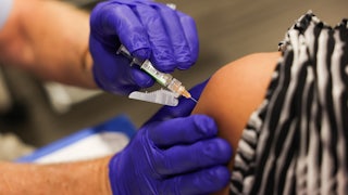 A person with purple gloves administers a vaccine dose in a syringe into another person's arm.