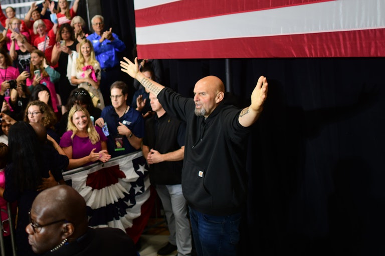 John Fetterman stands with his arms outstretched in front of an American flag and a crowd.