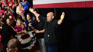 John Fetterman stands with his arms outstretched in front of an American flag and a crowd.