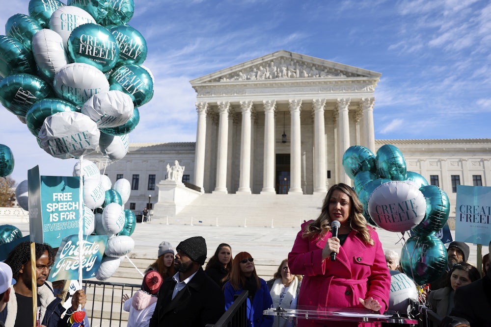 A woman in a pink coat speaks into a microphone at a podium in front of the Supreme Court. She is surrounded by supporters carrying signs and balloons that say CREATE FREELY and FREE SPEECH IS FOR EVERYONE.