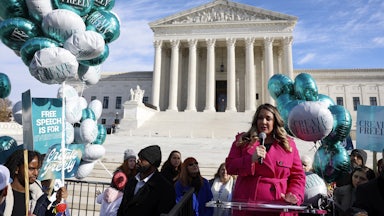 A woman in a pink coat speaks into a microphone at a podium in front of the Supreme Court. She is surrounded by supporters carrying signs and balloons that say CREATE FREELY and FREE SPEECH IS FOR EVERYONE.