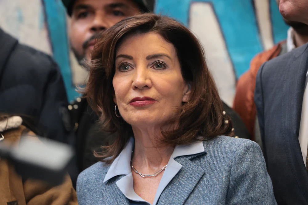 This picture shows a close-up of Kathy Hochul, wearing a light blue suit and blouse, with others semi-visible standing around her.