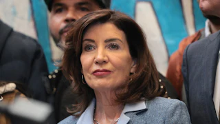This picture shows a close-up of Kathy Hochul, wearing a light blue suit and blouse, with others semi-visible standing around her.