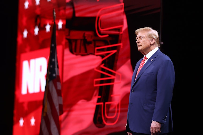 Trump stands in front of a red, black, and white background that reads "NRA" and "Trump." There is also a U.S. flag behind him.