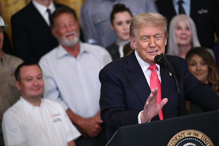Trump stands behind a lectern and in front of an audience and holds out a hand while he speaks. He's grinning