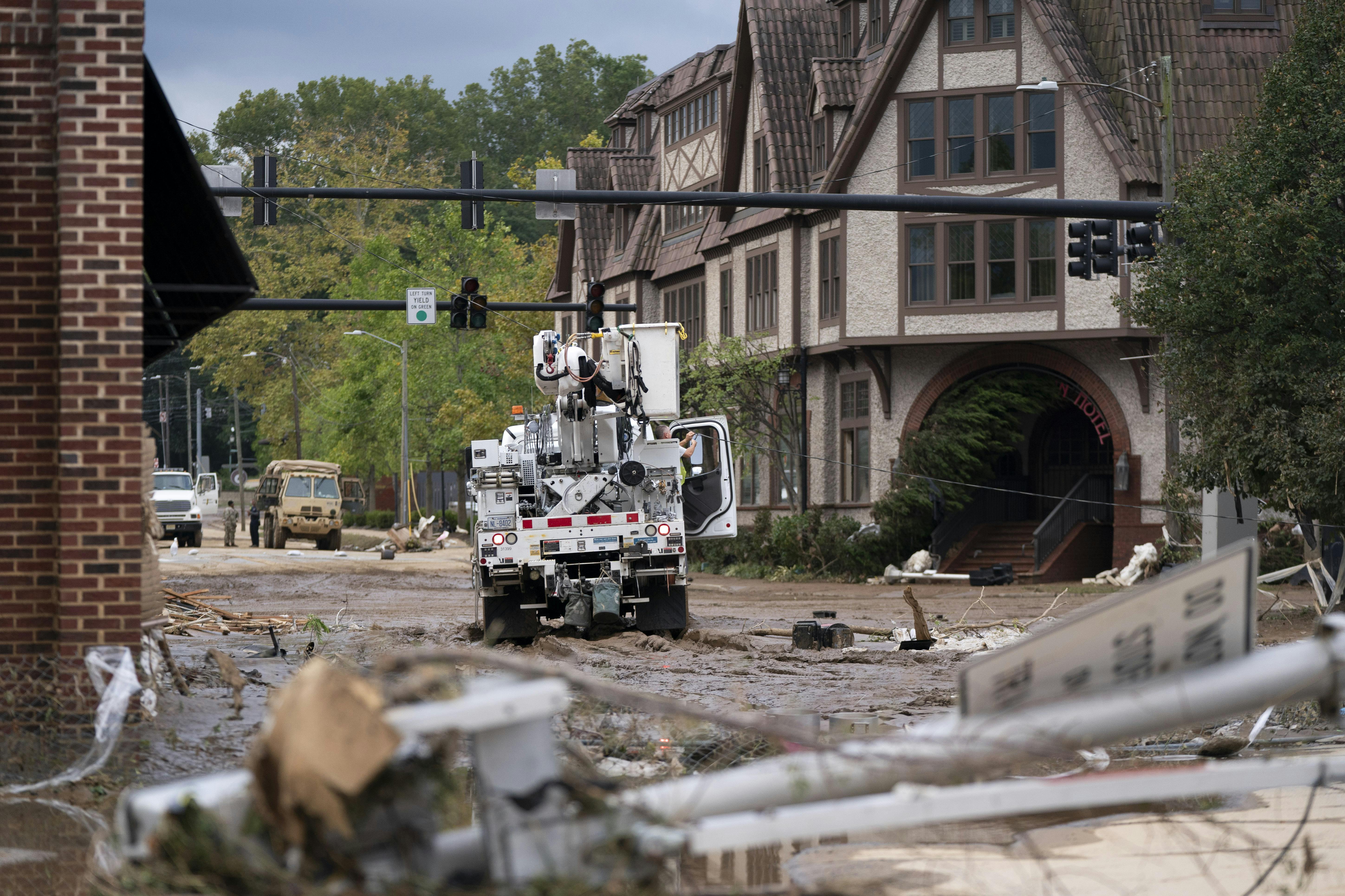 An electrical truck drives through a street full of mud, surrounded by buildings.