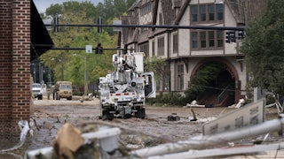 An electrical truck drives through a street full of mud, surrounded by buildings.
