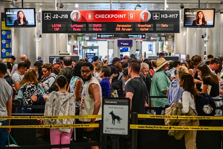 Screens play a video of Secretary of Homeland Security Kristi Noem as tons of travelers pass through Miami International Airport.