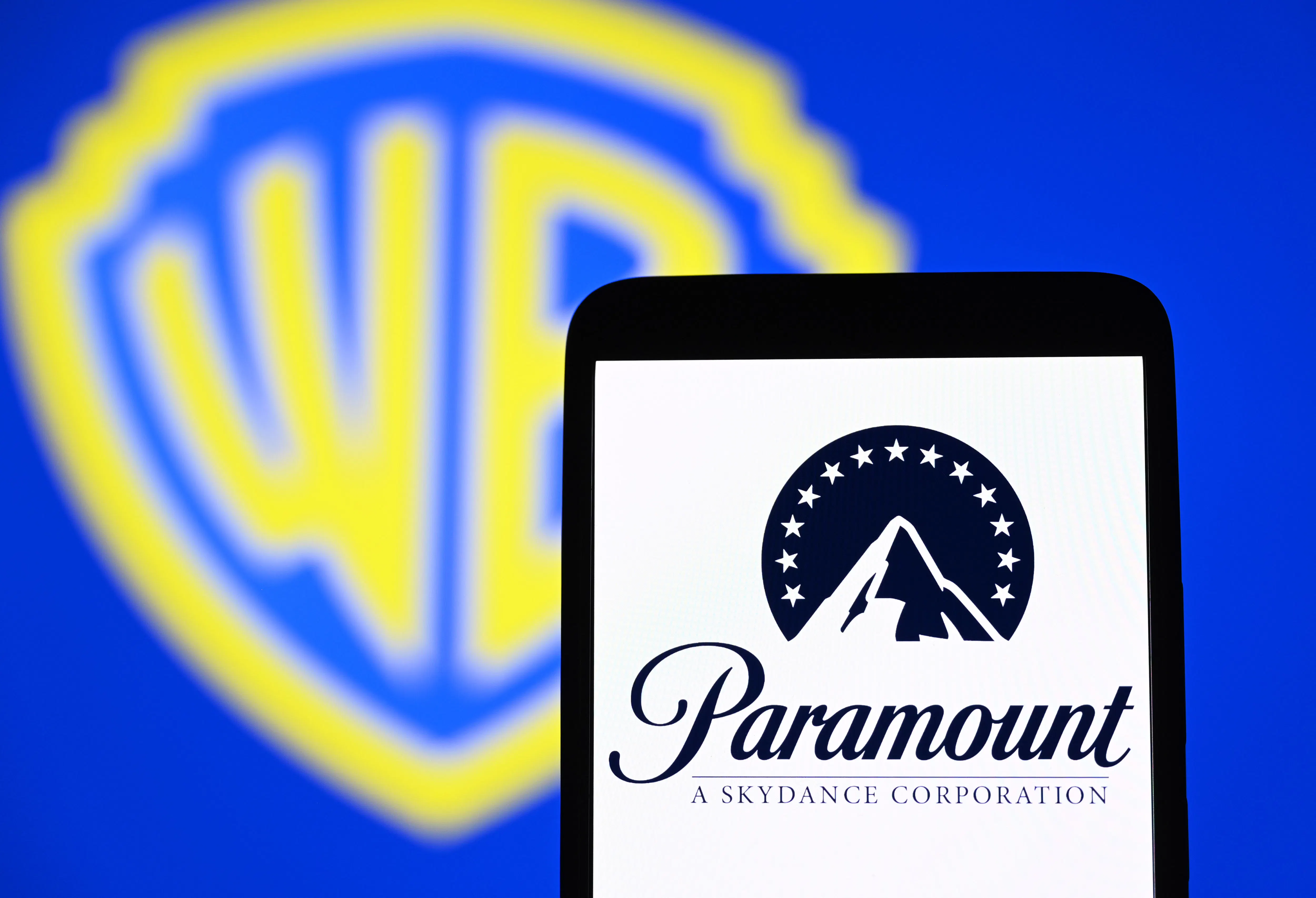 The Paramount Skydance Corporation logo is seen on a smartphone with the Warner Bros. logo in the background. 