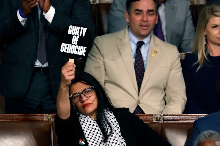 Rahida Tlaib wears a keffiyeh and holds up a sign that reads "Guilty of Genocide" while seated in the Capitol.